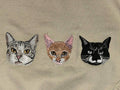 May include: Embroidered design featuring three cat faces. The cats are depicted in shades of gray, brown, and black, with detailed facial features and whiskers. The background is a solid beige color, providing a neutral backdrop for the cat portraits.