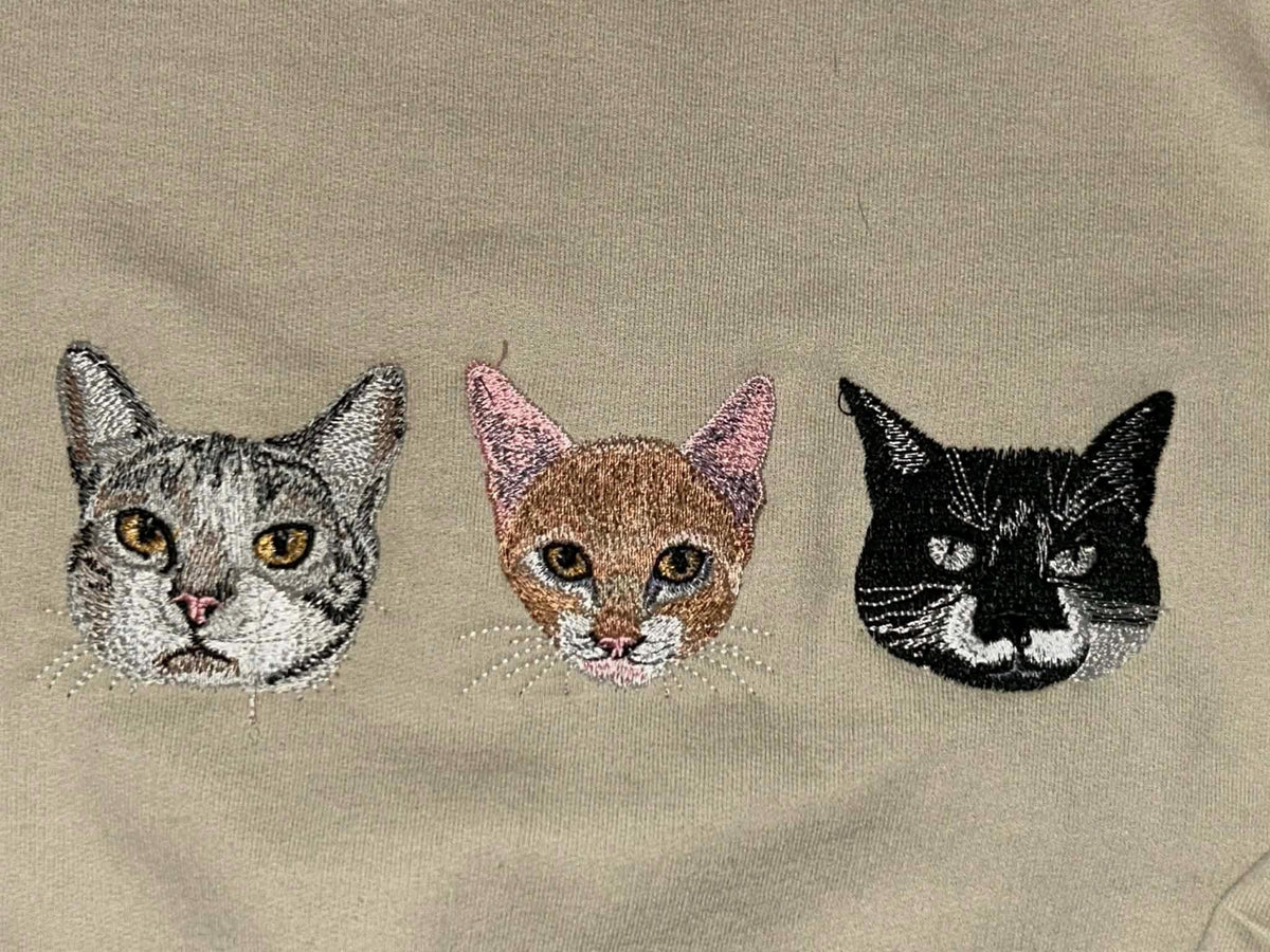 May include: Embroidered design featuring three cat faces. The cats are depicted in shades of gray, brown, and black, with detailed facial features and whiskers. The background is a solid beige color, providing a neutral backdrop for the cat portraits.