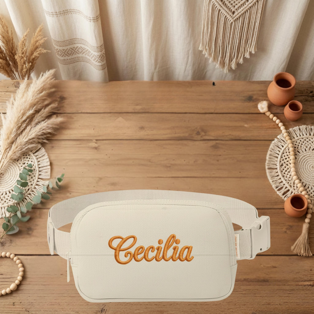 A cream-colored fabric belt bag with the name 'Cecilia' embroidered in orange cursive script on the front, displayed on a rustic wooden table surrounded by boho-style decorative props.